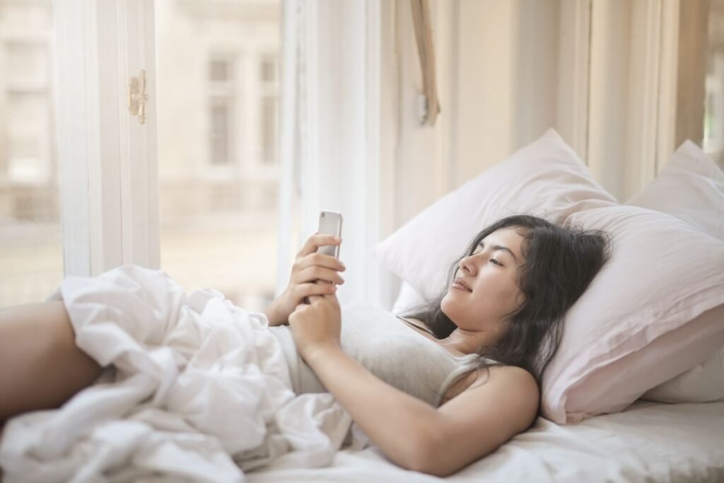 Young woman lying in bed using her smartphone in soft morning light